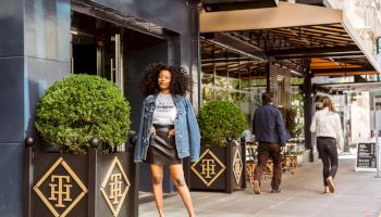 Woman posing on sidewalk, storefront behind, people walking, decorative plants in front.