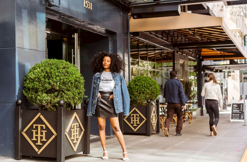 Woman posing on sidewalk, storefront behind, people walking, decorative plants in front.