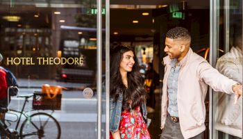 A man and woman are exiting Hotel Theodore with smiles, while he holds the door open and a cyclist rides by outside.