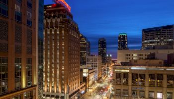 This image shows a cityscape at dusk with tall buildings, neon signs, and busy streets illuminated by streetlights. Buildings display 