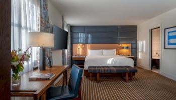 The image shows a modern hotel room with a king-sized bed, a desk and chair, a TV mounted on the wall, and an adjacent bathroom.