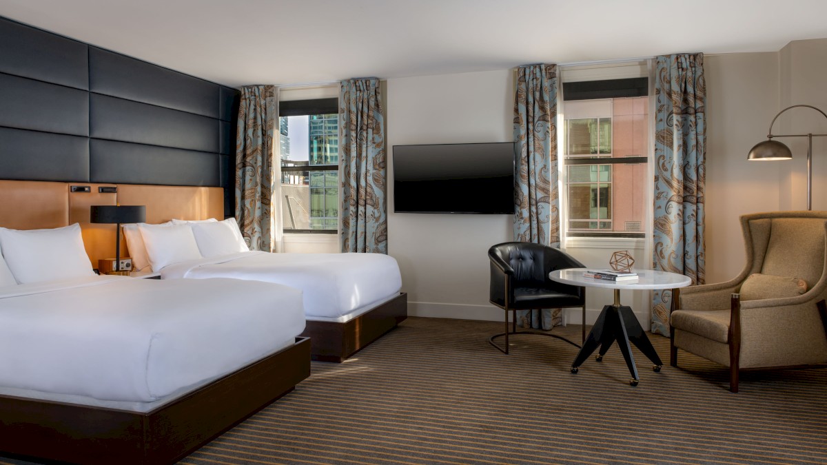 A modern hotel room with two double beds, a wall-mounted TV, a round table, chairs, and a floor lamp near the window.