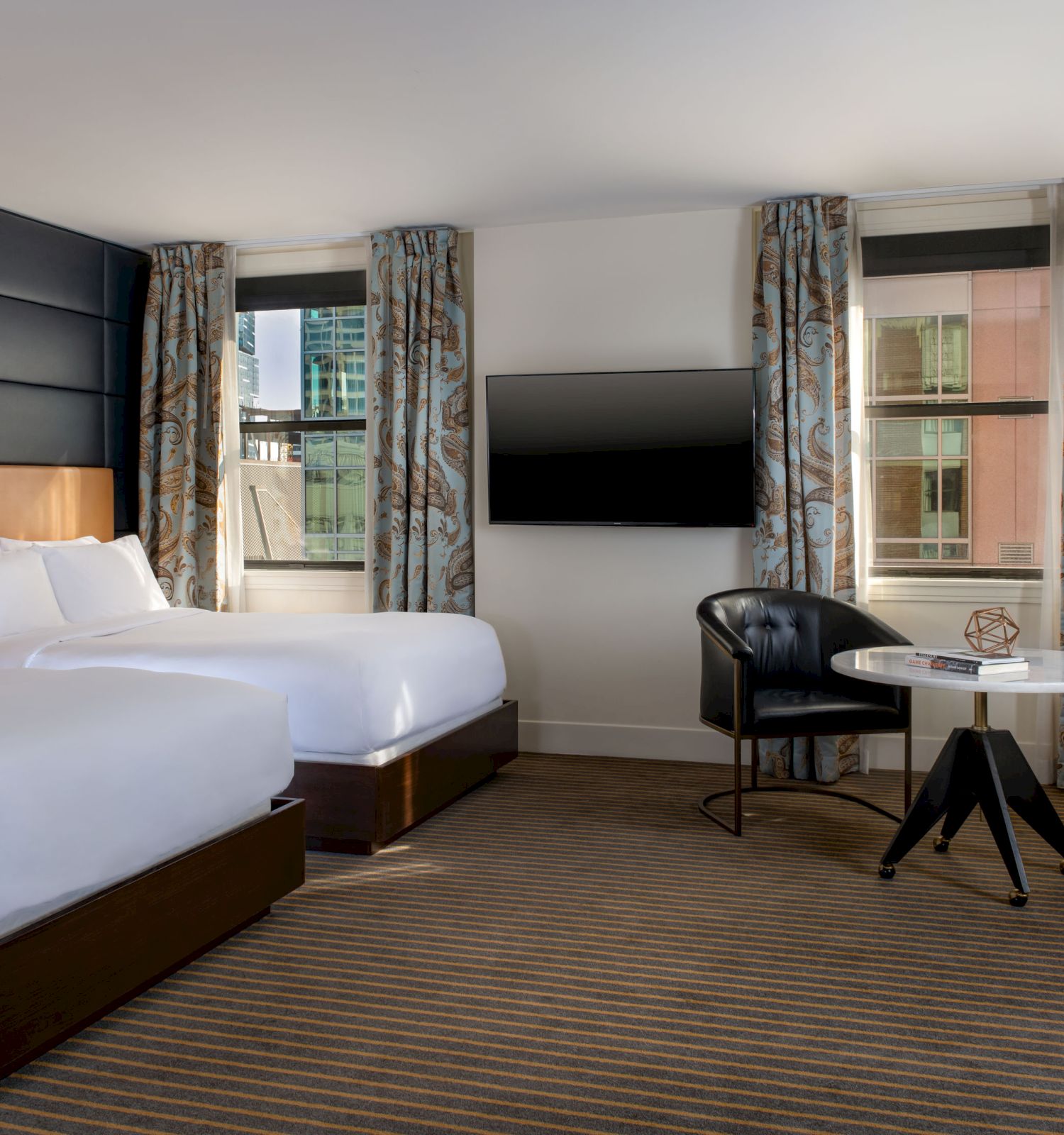 A modern hotel room with two double beds, a wall-mounted TV, a round table, chairs, and a floor lamp near the window.