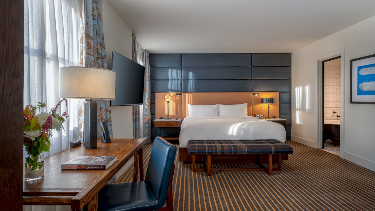 A well-lit hotel room with a king-size bed, desk and chair, wall-mounted TV, striped carpet, and a doorway leading to the bathroom.