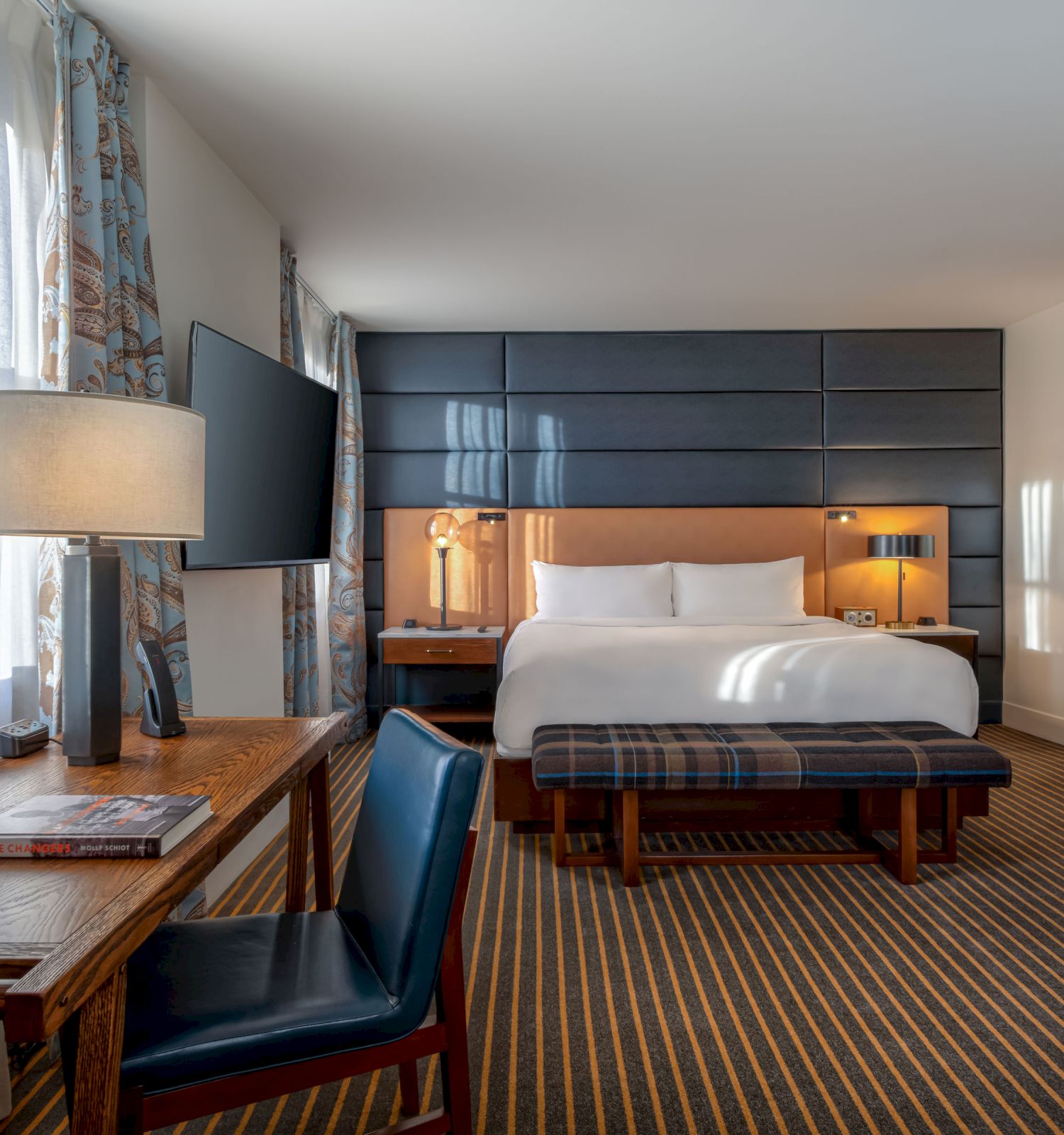 A well-lit hotel room with a king-size bed, desk and chair, wall-mounted TV, striped carpet, and a doorway leading to the bathroom.