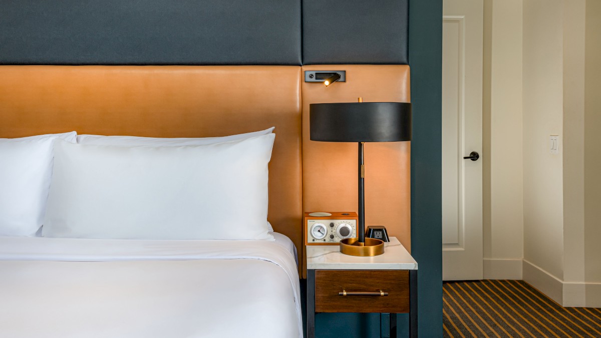 A neatly made bed with white linens is next to a wooden nightstand holding a lamp, phone, and small tray. A doorway is visible to the right.