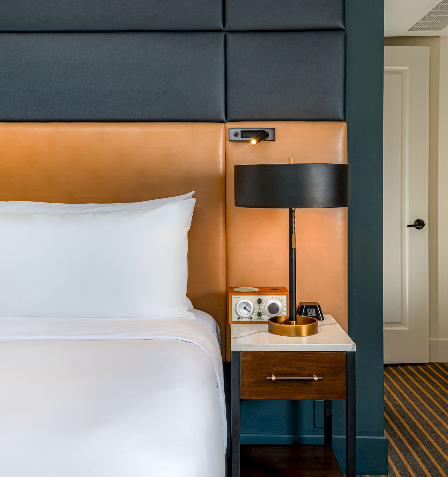 A neatly made bed with white linens is next to a wooden nightstand holding a lamp, phone, and small tray. A doorway is visible to the right.