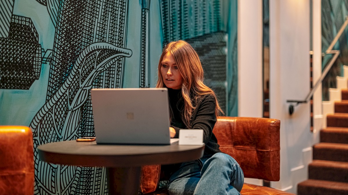 A person is seated at a table with a laptop in front of a mural, working or browsing. The setting appears to be indoors with a staircase nearby.