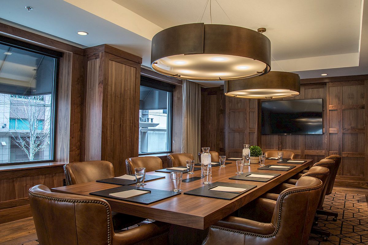 A modern conference room with a large wooden table, leather chairs, large hanging lights, windows, and a wall-mounted TV, ending the sentence.