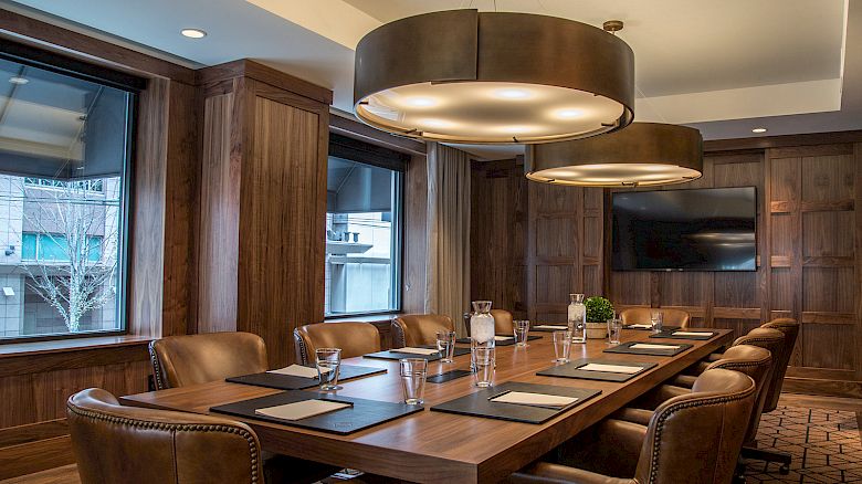 A modern conference room with a large wooden table, leather chairs, large hanging lights, windows, and a wall-mounted TV, ending the sentence.