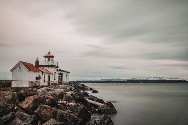 A tranquil lighthouse sits on a rocky shore as calm water stretches out, clouds drift softly overhead, and distant land anchors the horizon.