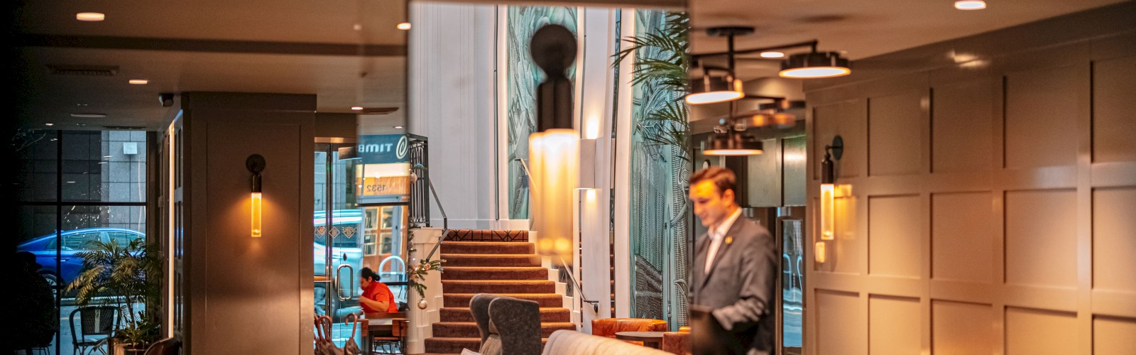 A man in a suit stands in a warmly lit lobby with a mix of modern and classic decor, including cozy seating and wall sconces.