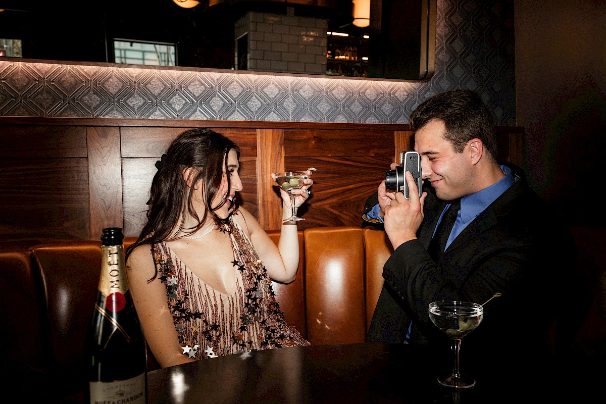 A woman holds a drink in a bar while a man photographs her. A bottle and cocktail are on the table in front of them.