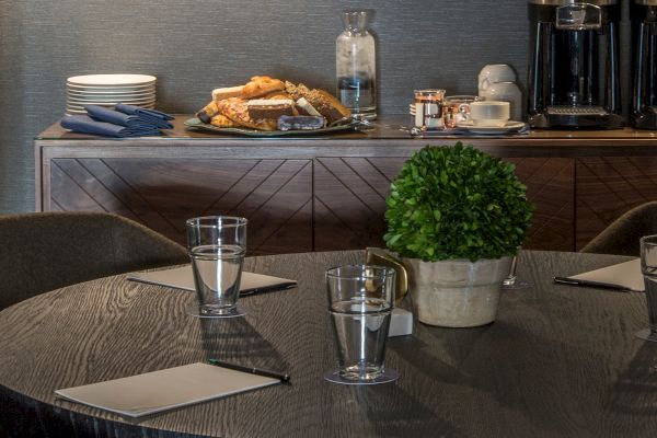 A round table with glasses, notepads, and a potted plant; behind it, a buffet with plates, pastries, and a coffee station ending the sentence.