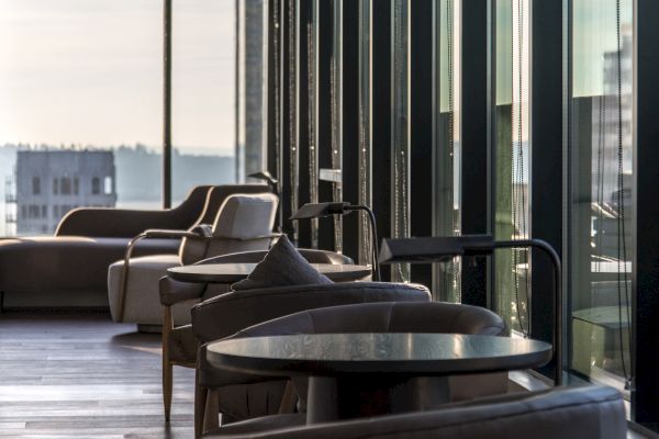 A modern lounge area with large windows, natural light, comfortable chairs, and tables, overlooking a cityscape.
