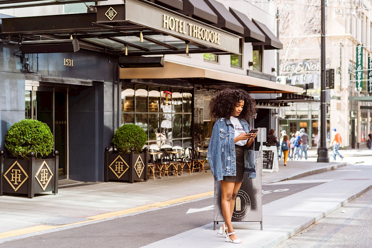 A woman stands outside Hotel Theodore, reading a book near a street corner. The hotel and some pedestrians are visible in the background.
