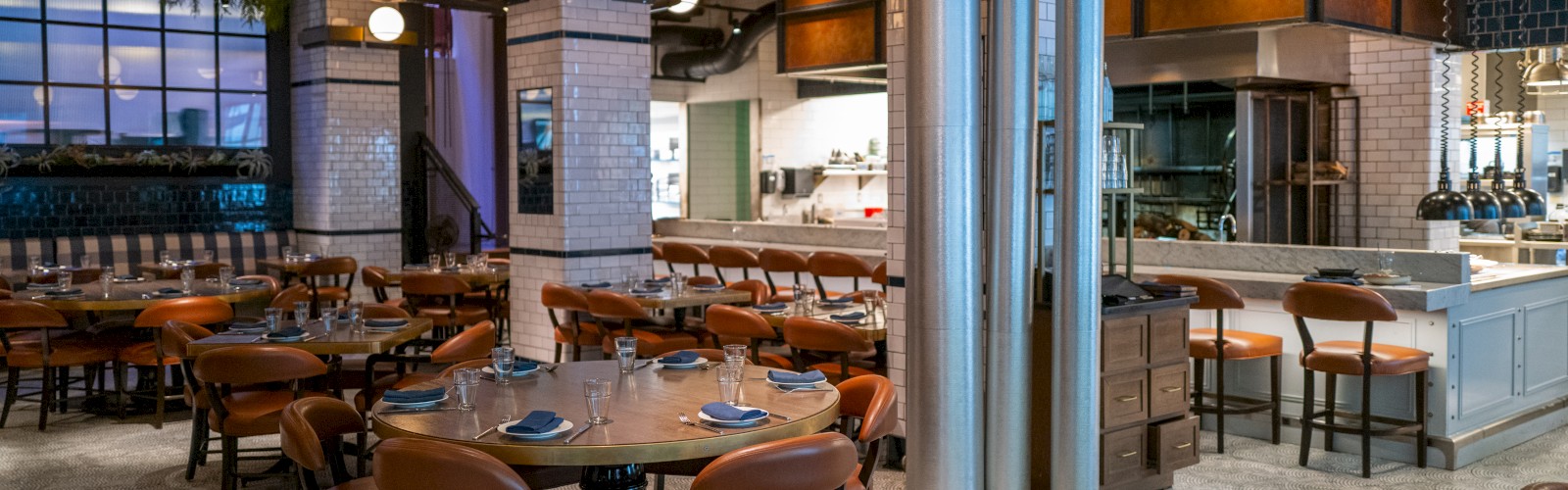 The image shows a modern, spacious restaurant with round tables and brown leather chairs, industrial decor, exposed ducts, and globe light fixtures.