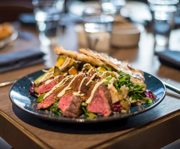 A plate of sliced steak with greens, roasted vegetables, and drizzled sauce is on a table set with glasses and napkins.