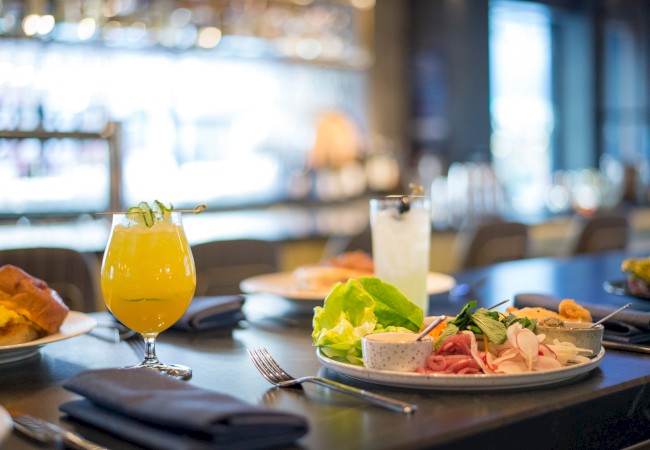 A dining table with various dishes and drinks, including a yellow cocktail and a salad, set in a modern restaurant.