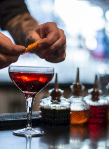 A bartender is garnishing a cocktail in a glass, with various containers of ingredients in the background, ending the sentence.