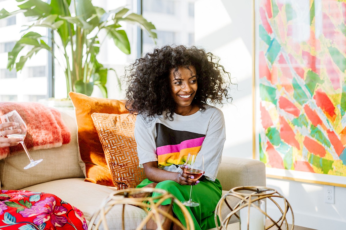 A person with curly hair is sitting on a couch, holding a glass, with colorful decor and plants in the background.