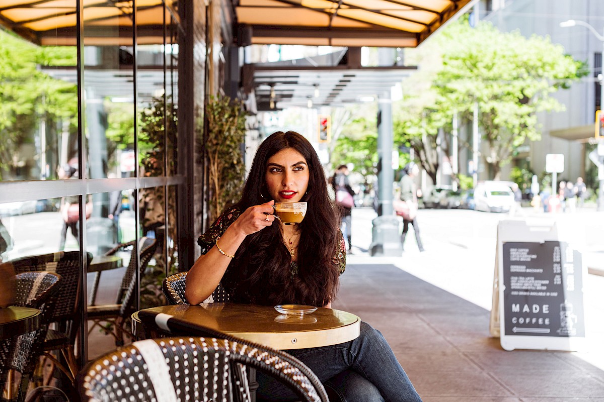 A woman is sitting at an outdoor café table, sipping a drink, with the city street in the background under a canopy on a sunny day, ending the sentence.