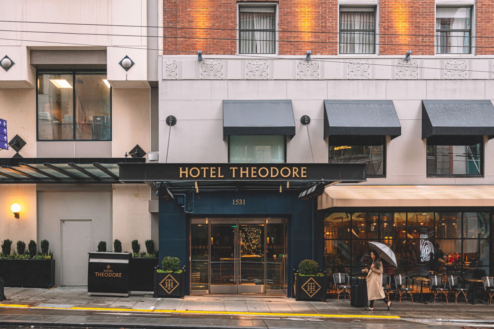 The image shows the entrance of Hotel Theodore with a person holding an umbrella walking by. The building features a mix of modern and classic design.