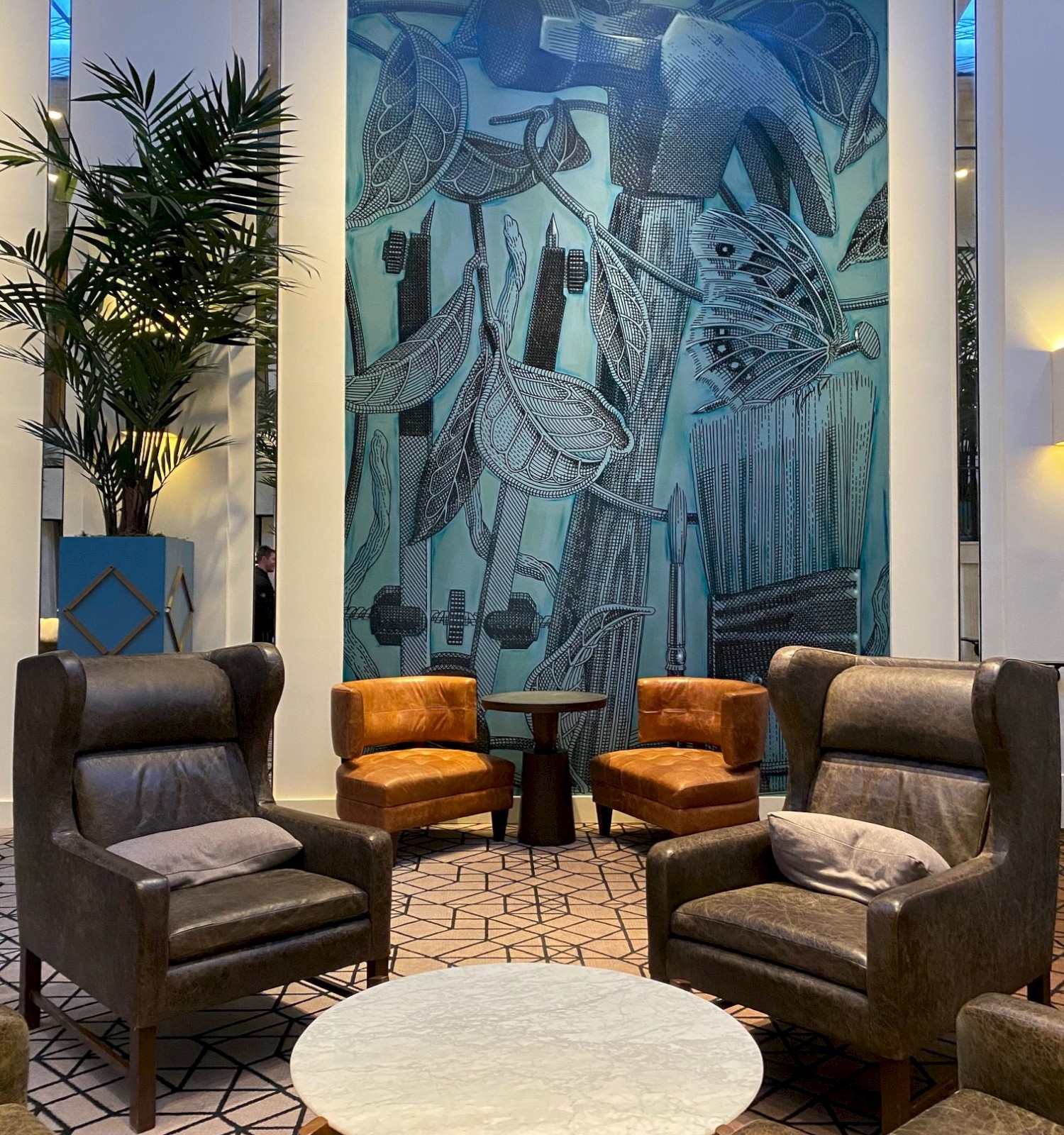 A modern lounge area with chairs, a round table, and a large blue abstract painting on the wall in the background, complemented by tall indoor plants.