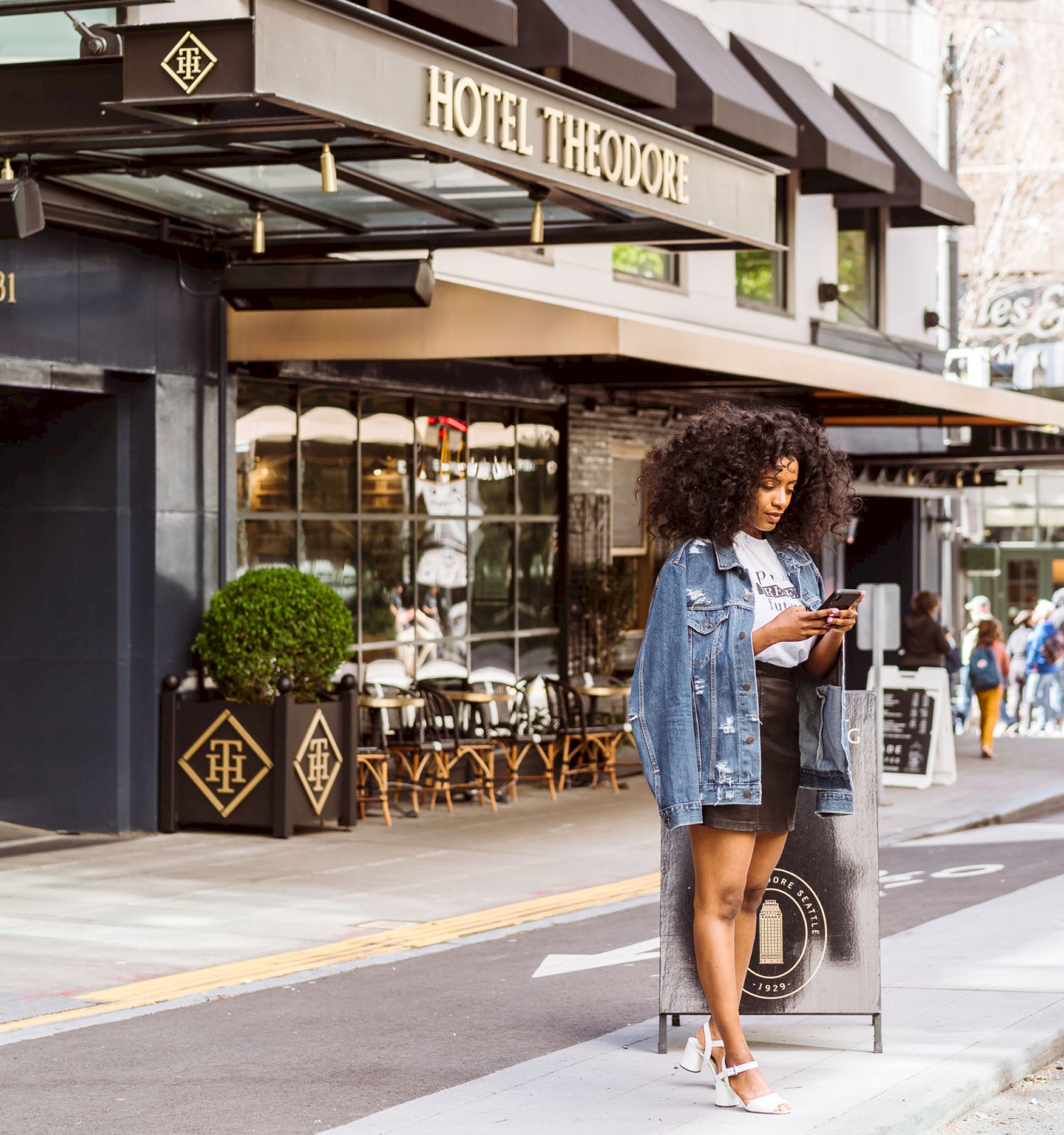 A person is standing outside Hotel Theodore reading or using a tablet, with people walking by in the background and trees lining the street.