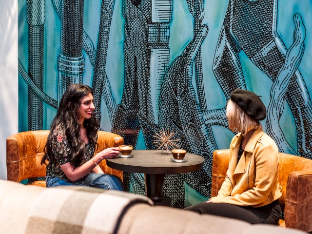 Two people are sitting at a round table with cups of coffee, engaging in conversation in a cozy room with modern decor and vibrant artwork.
