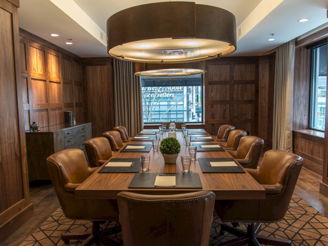 A conference room with a large wooden table, leather chairs, elegant lighting, and a window view, set for a meeting with notepads and pens.