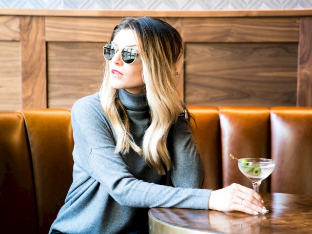 A person wearing sunglasses and a gray sweater sits at a table, holding a martini with olives in a stylish, wood-paneled setting.
