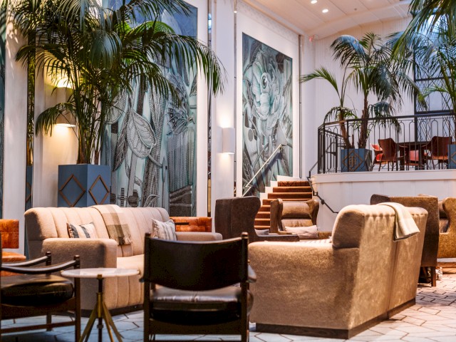A stylish lounge area with comfortable seating, tall indoor plants, and large wall murals, featuring a staircase to an upper dining or seating area.