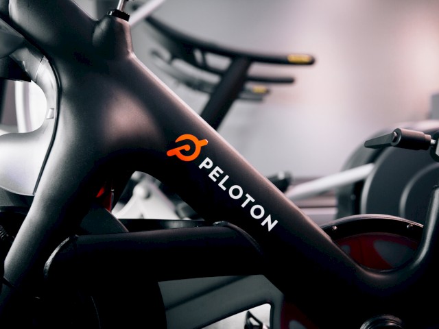 The image shows a close-up of a Peloton exercise bike, focusing on the Peloton logo and branding on the bike frame in an indoor setting.
