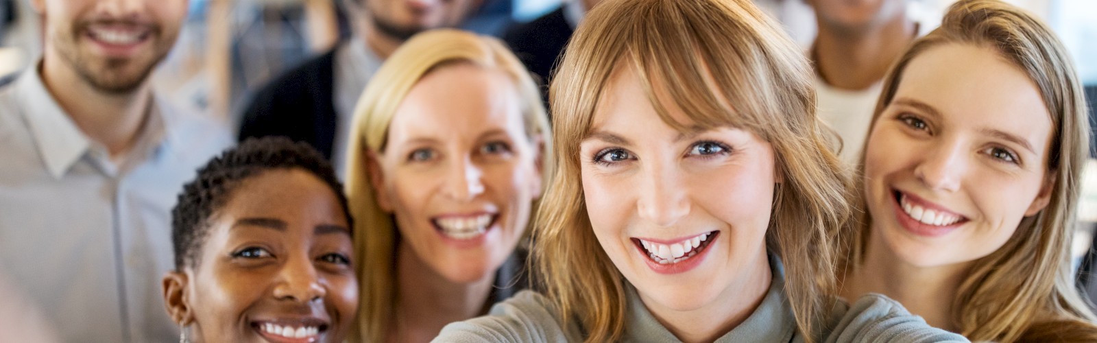 A group of eight people are taking a cheerful selfie, smiling and standing close together in what appears to be an indoor setting.