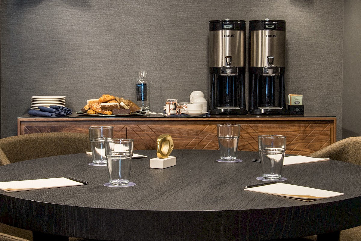 A round table with four chairs, glasses, and napkins in the foreground; coffee dispensers, pastries, and cups on a sideboard in the background.