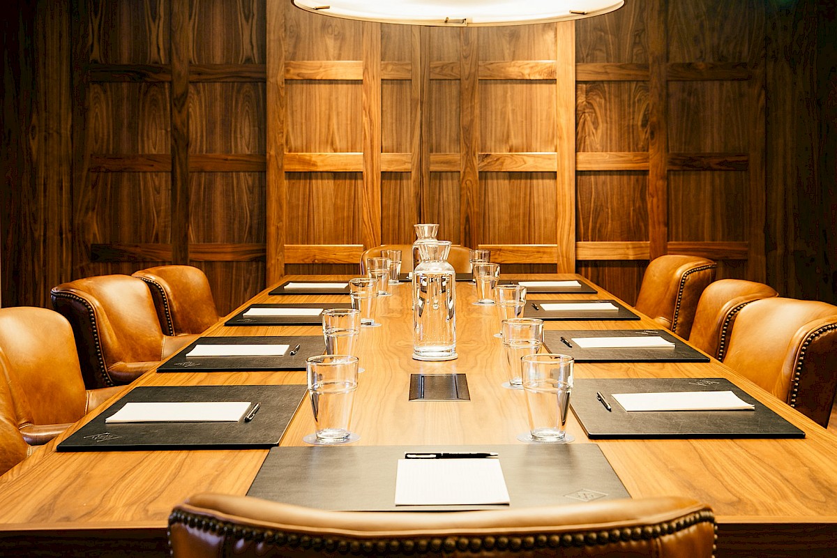A boardroom setup with a long wooden table, leather chairs, notepads, glasses, and a central water pitcher, illuminated by overhead light.