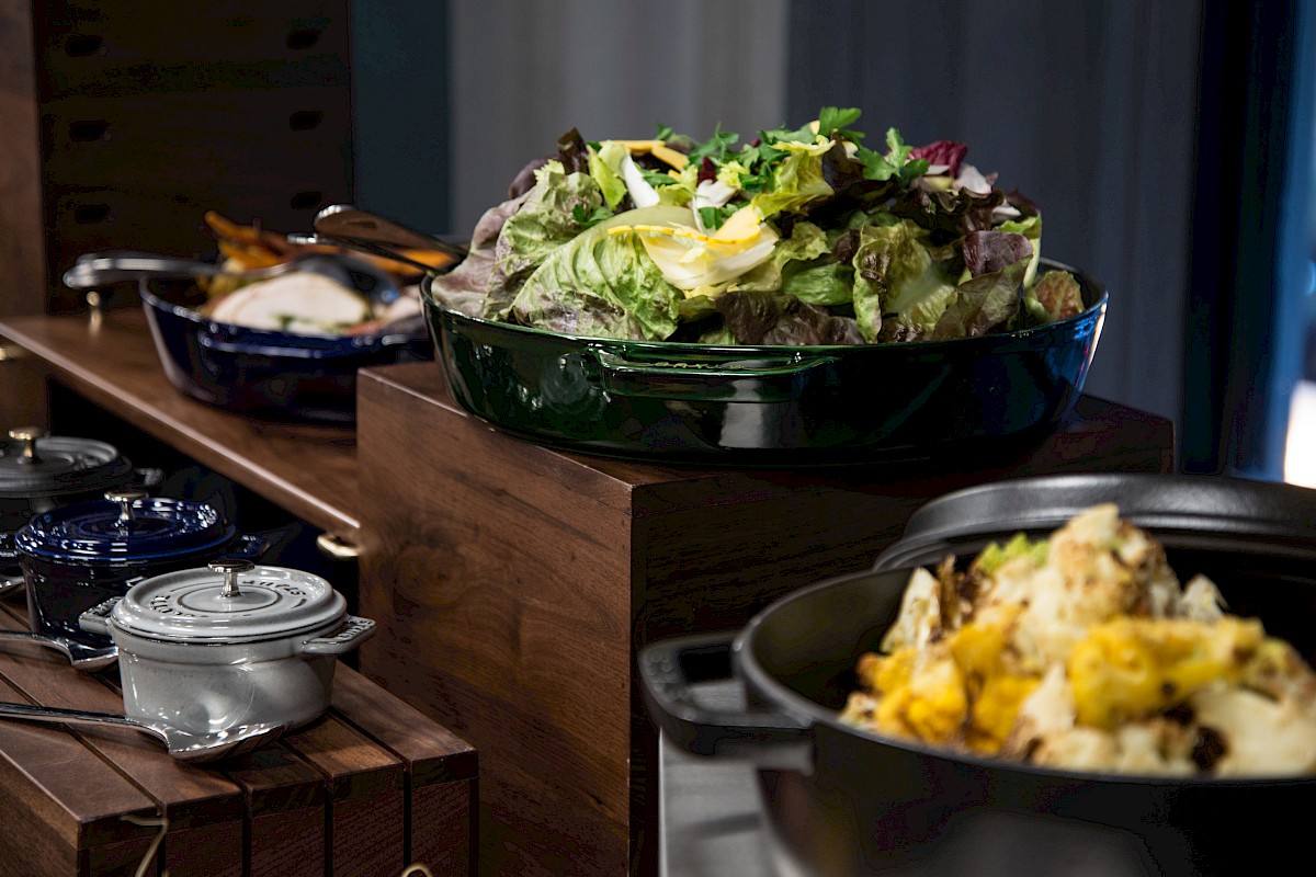The image shows a buffet with dishes including a green bowl of salad and a black pot with a vegetable dish.