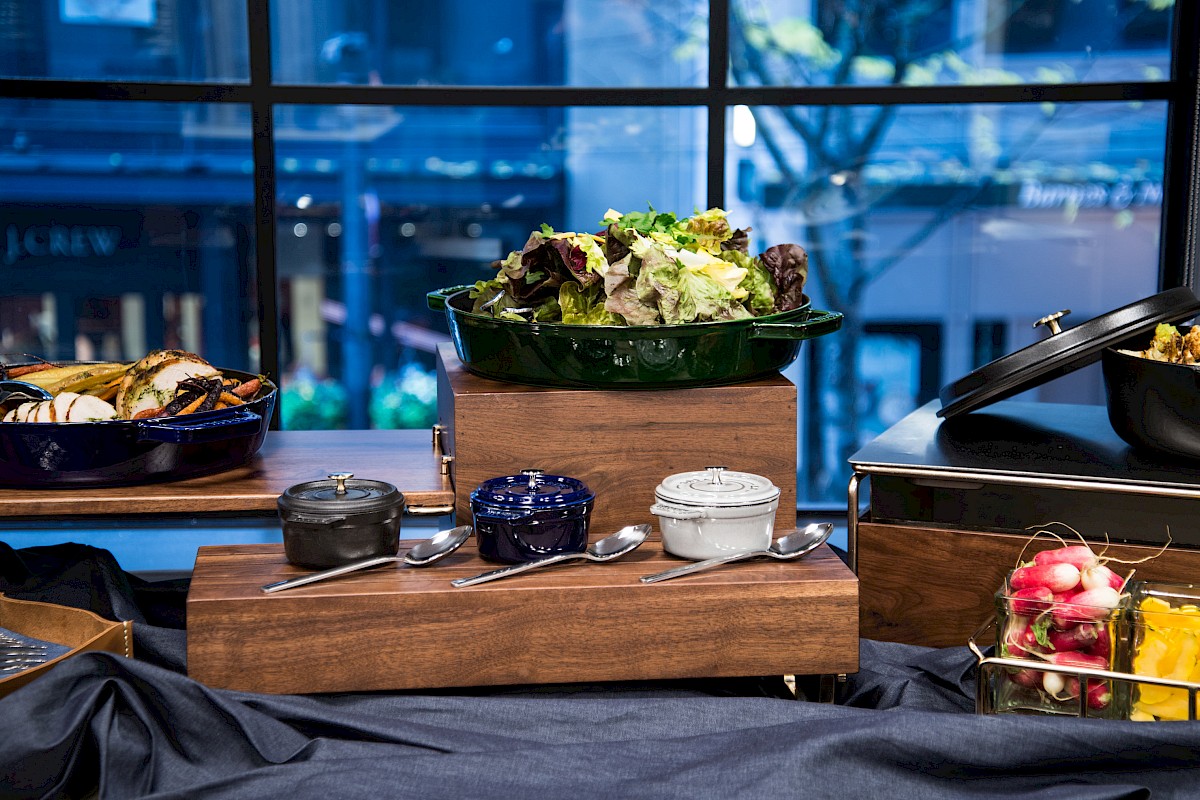 A buffet spread featuring a salad bowl, condiments in small jars, and assorted foods in serving dishes, all placed on a wooden table.