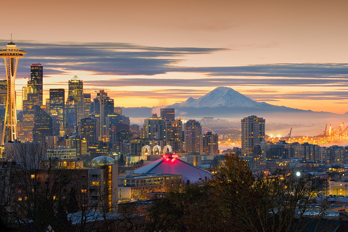 A cityscape at dusk featuring a skyline with a prominent tower, mountains in the background, and colorful lights throughout.