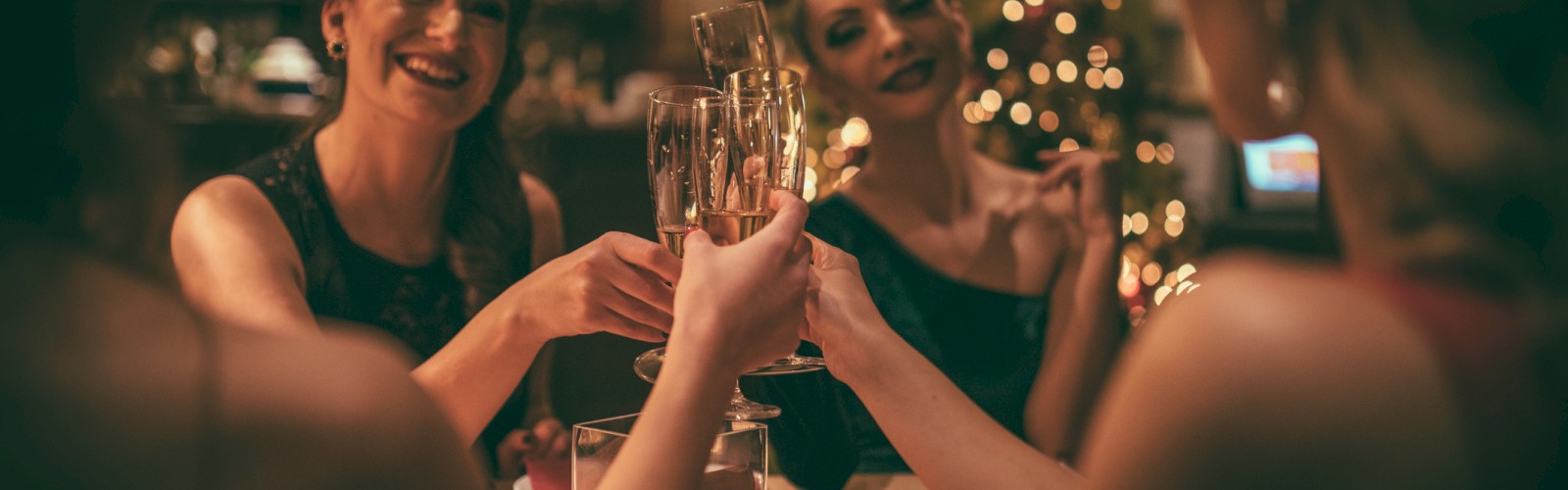 A group of women are raising glasses in a toast, sitting at a table in a warmly lit, festively decorated setting.