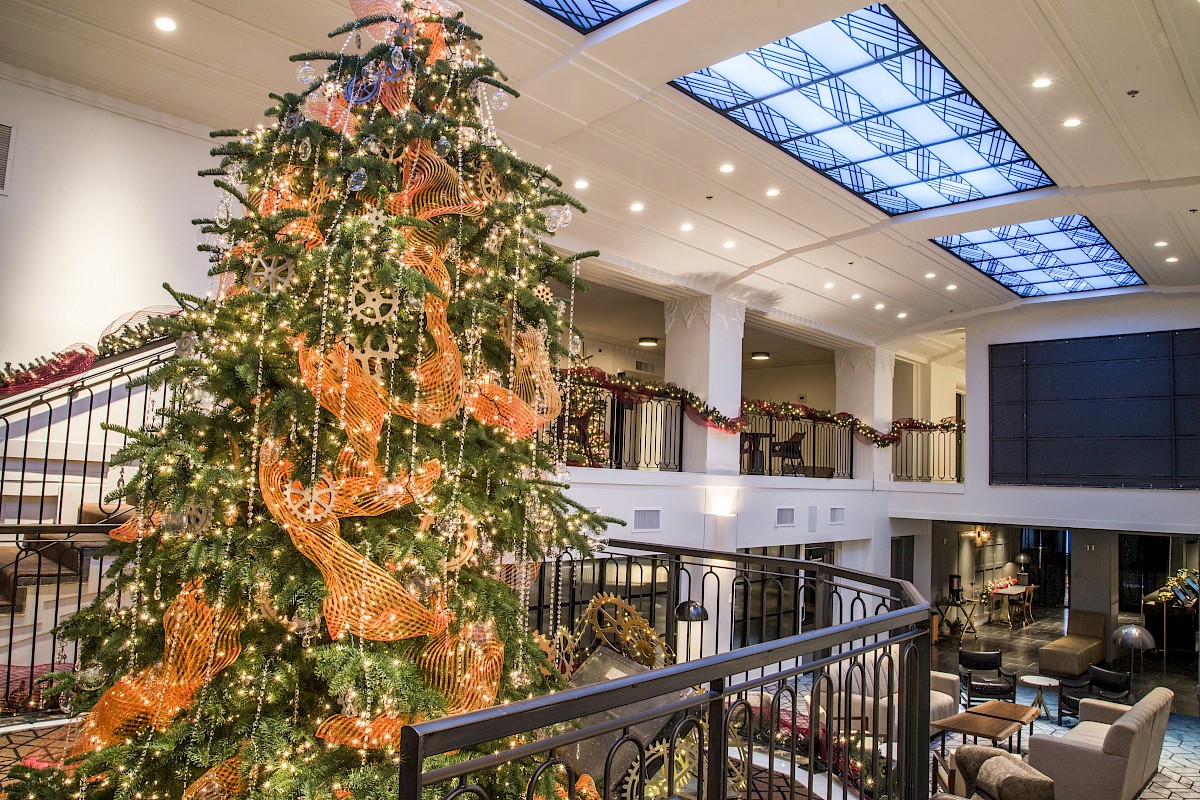 The image shows a decorated Christmas tree with orange ribbons and lights in an elegant, spacious indoor area with a glass ceiling and seating.
