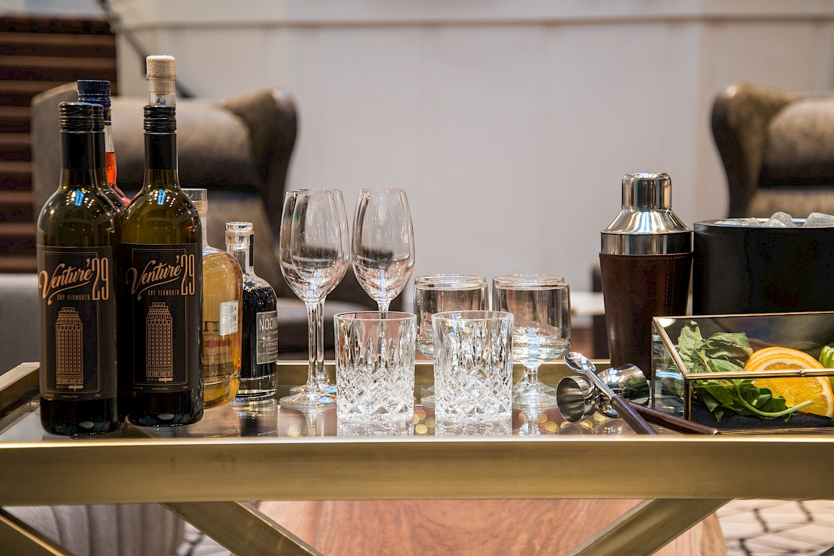A bar cart with liquor bottles, wine glasses, tumblers, a cocktail shaker, ice bucket with ice, citrus slices, and fresh herbs.