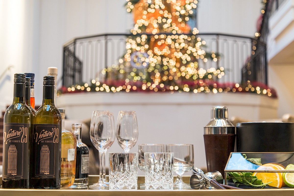 The image shows a festive setup with a Christmas tree in the background, bottles of liquor, glasses, and cocktail-making supplies in the foreground.