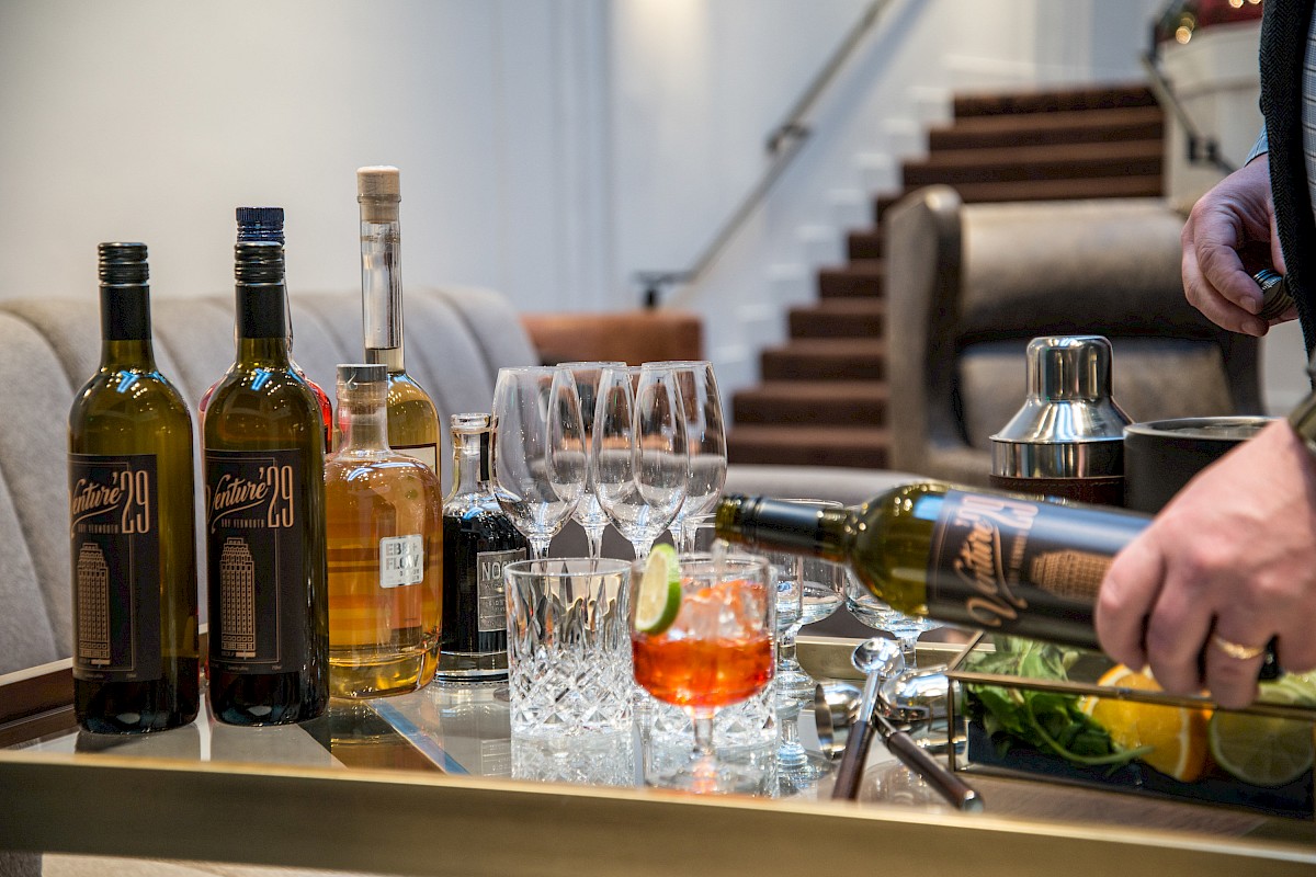 A table with various liquor bottles, glasses, and a cocktail being prepared with mixers, and garnishes. There are stairs and chairs in the background.