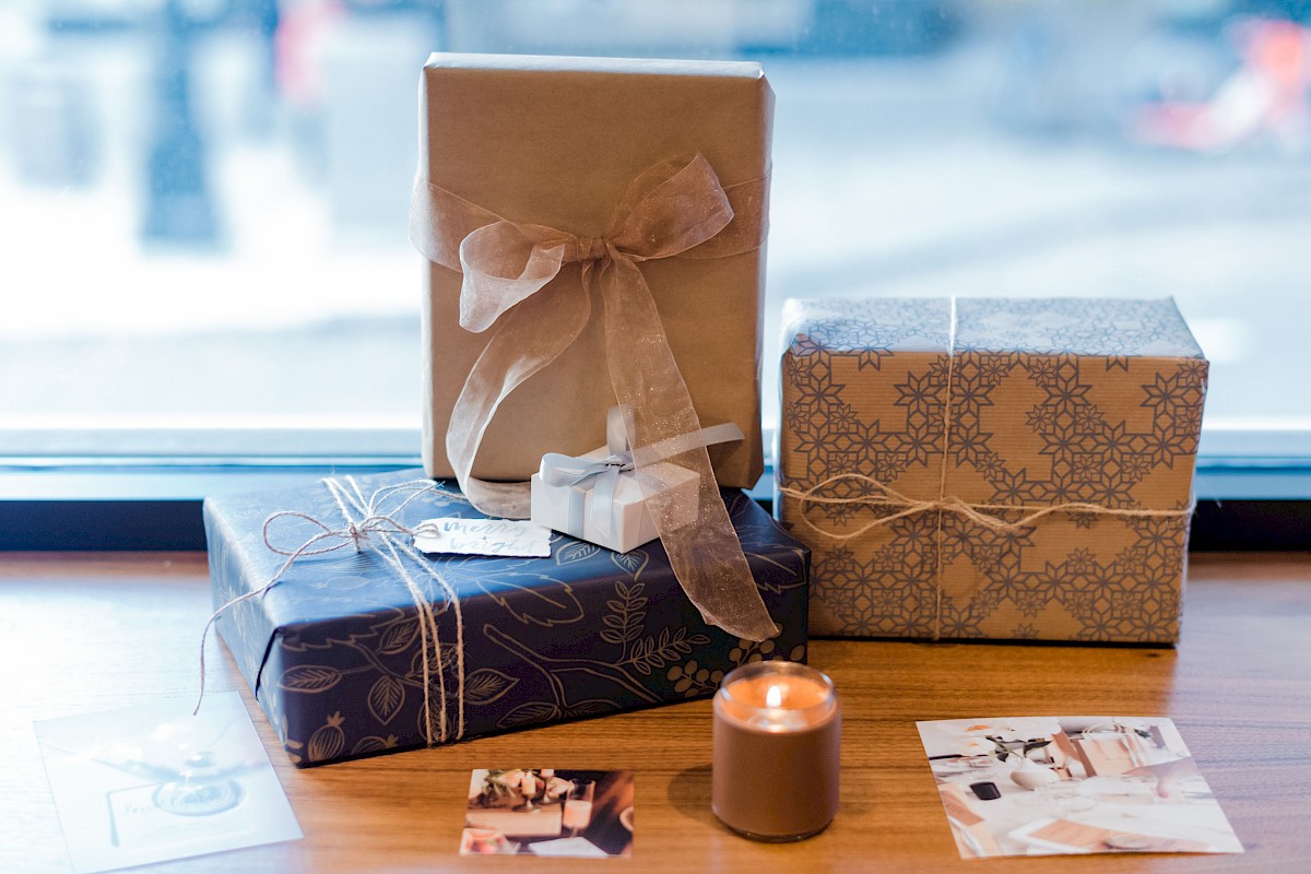 The image shows three wrapped gifts with ribbons, a lit candle, and a few photographs on a wooden surface near a window.