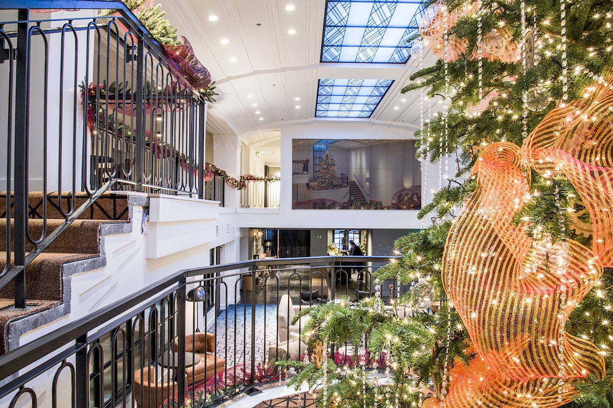 The image shows a decorated interior space with a large Christmas tree adorned with lights, a staircase, and railings leading to another level.