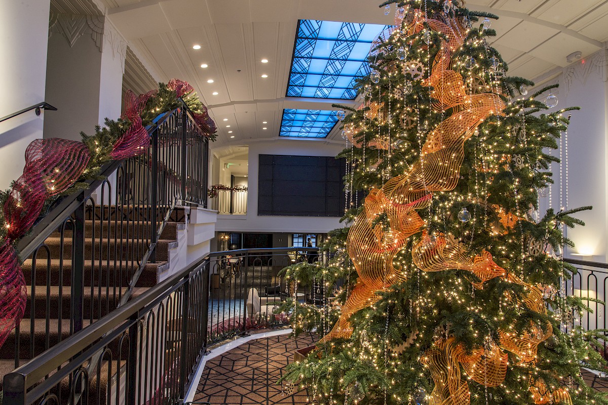 A large Christmas tree decorated with lights and ribbons is near a staircase adorned with festive garlands in a spacious, well-lit room.