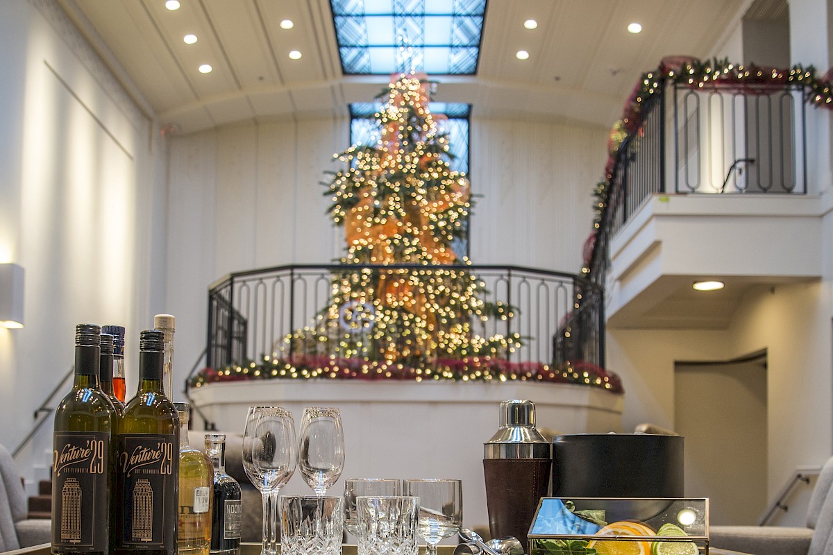 A festive room with a decorated Christmas tree, bottles, glasses, and a cocktail setup on the table in the foreground.