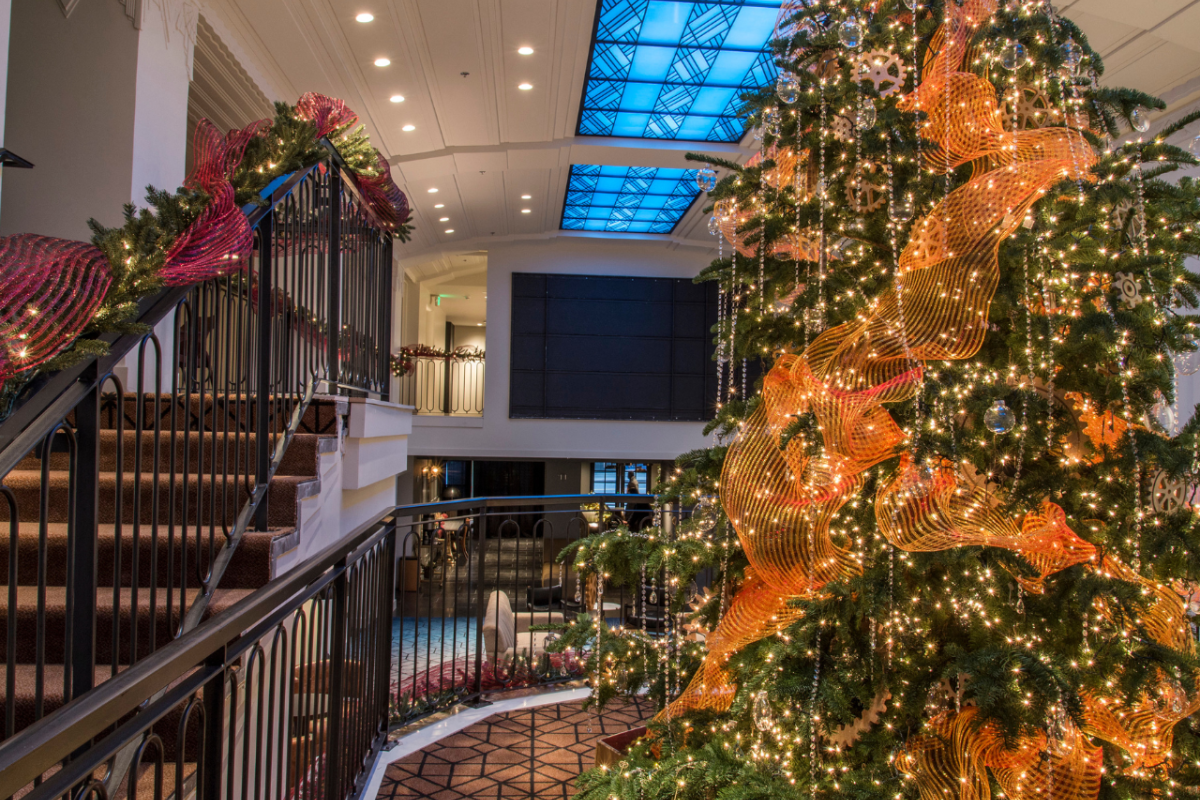 The image shows a beautifully decorated indoor Christmas tree next to a staircase, adorned with lights and festive ornaments.